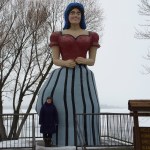 a woman standing next to a very tall statue of a woman in a red shirt and blue-and-black striped dress