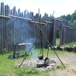 cast-iron pot hanging over a small fire, with a table nearby, a tall wooden fence in the background