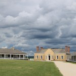 A house with four chimneys, in between two long white buildings, with gray clouds